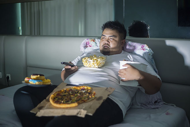Unhealthy lifestyle concept: Asian obese man eating junk foods while watching TV in bed before sleep