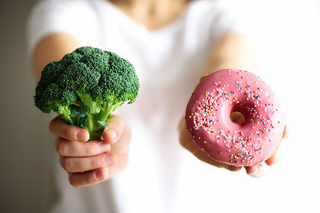 Young woman in white T-shirt choosing between broccoli or junk food, donut. Healthy clean detox eating concept. Vegetarian, vegan, raw concept. Copy space.