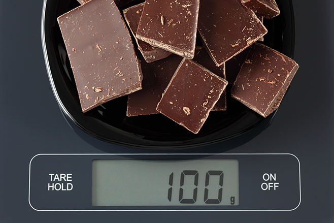 Broken dark chocolate in a black plate on digital scale displaying 100 gram.