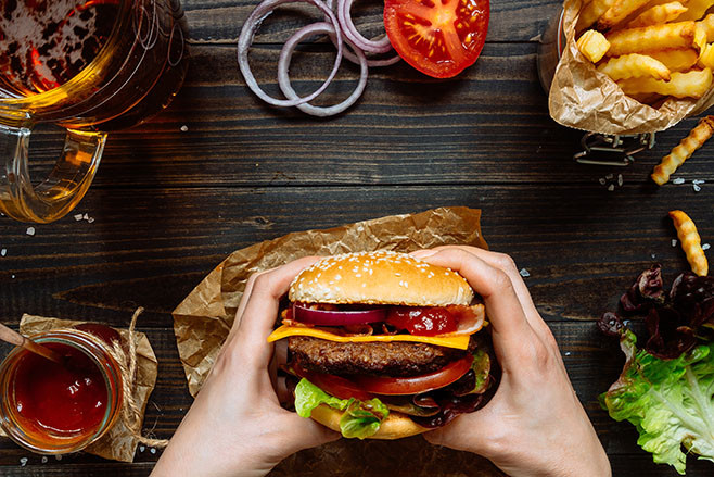 Hands holding fresh delicious burgers with french fries, sauce and beer on the wooden table top view.