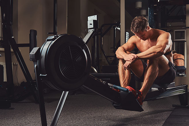 A muscular shirtless athlete resting after a hard workout while sits on the rowing machine in the gym.