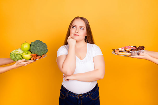 Body care bodycare concept. Confused thoughtful hesitating fatty woman can't make a choice between healthy natural products and sugary tasty biscuits, isolated on bright yellow background