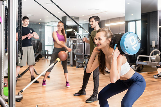 Young determined fit woman doing squats exercise, while holding on the back of her neck a heavy sandbag during group circuit functional training at the gym