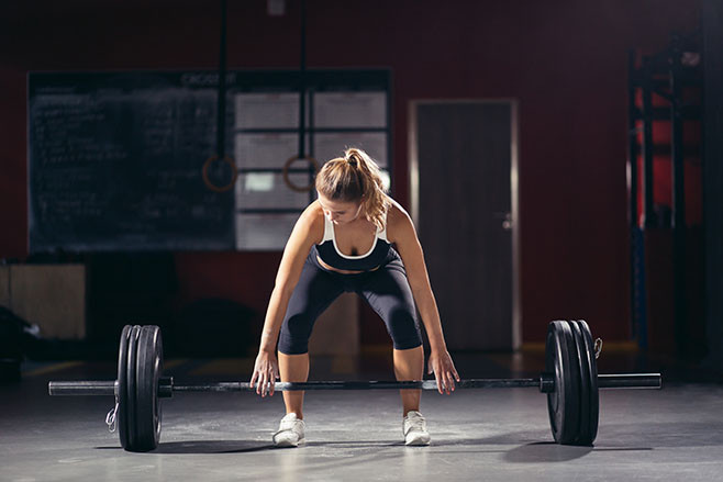 Intense workout in dark gym: young athletic woman doing deadlift with barbell on standing position, legs at shoulder level.