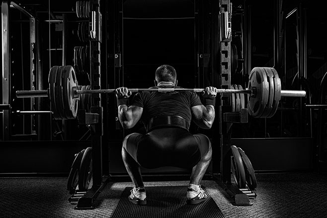 Professional athlete sits with a barbell on his shoulders and prepares to stand with her. View from the back.